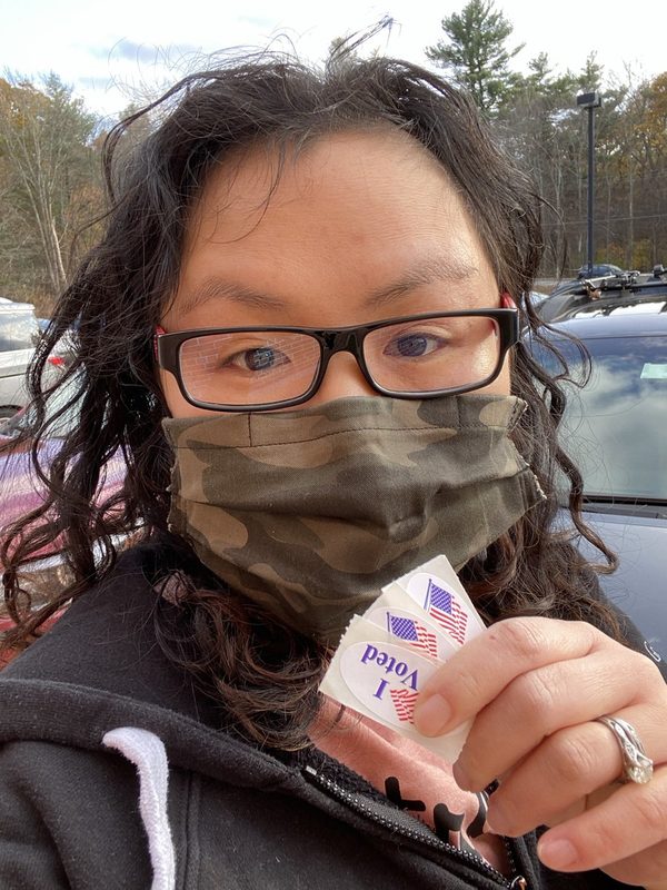 Annie, masked, holding a fan of "I Voted" stickers in a parking lot